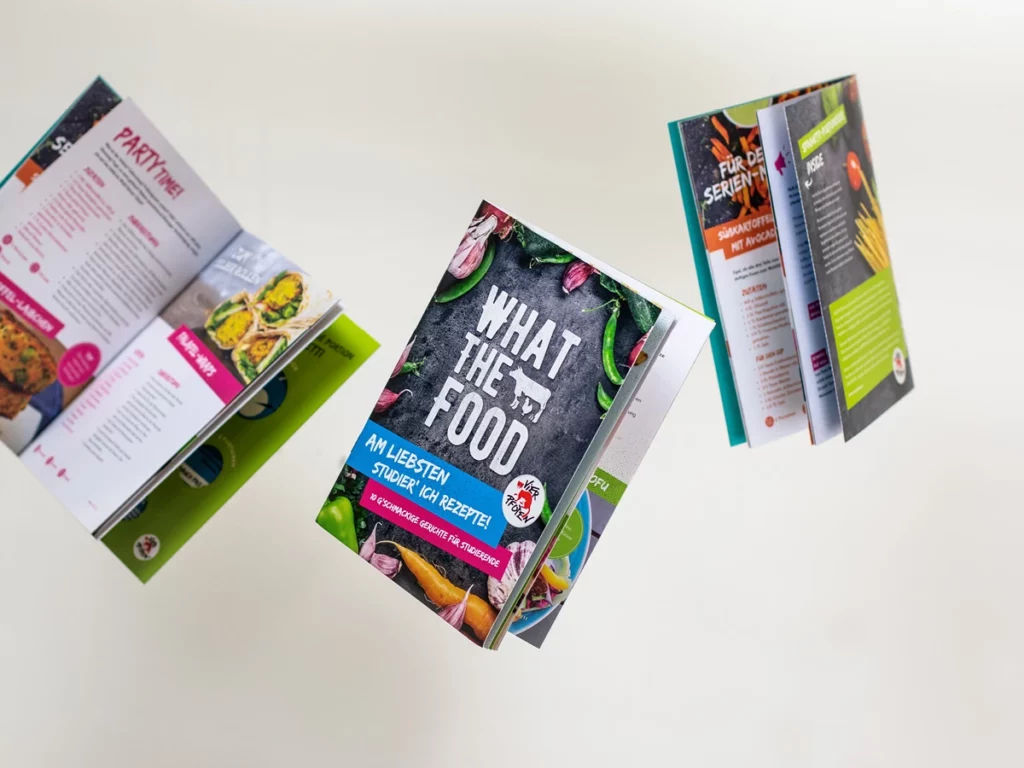 Mehrere Exemplare der Vier-Pfoten-Broschüre „What the Food“ schweben aufgefächert im Raum, aufmerksamkeitsstarkes Grafikdesign als Teil professioneller Werbung einer Werbeagentur in Wien.